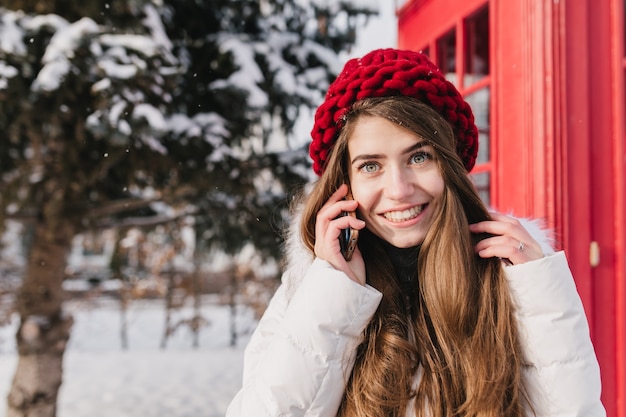 Elegante retrato británico de increíble joven con cabello largo morena con sombrero rojo hablando por teléfono en la calle llena de nieve. Disfrutando del frío invierno, buen humor. Lugar para el texto.