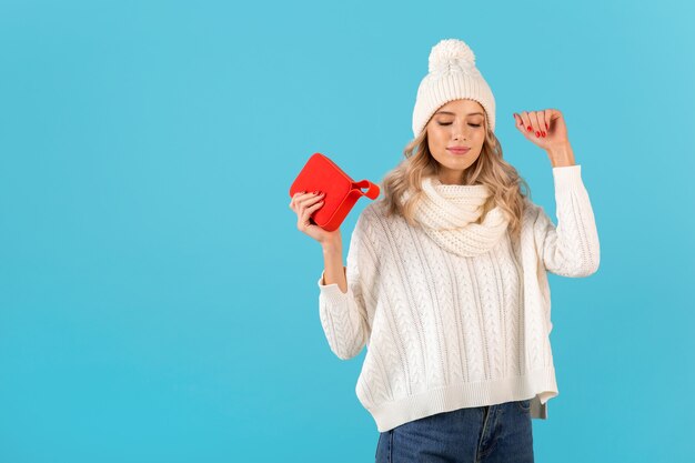 Elegante rubia sonriente hermosa joven sosteniendo altavoz inalámbrico escuchando música feliz bailando vistiendo suéter blanco y sombrero de punto estilo de invierno posando aislado en la pared azul