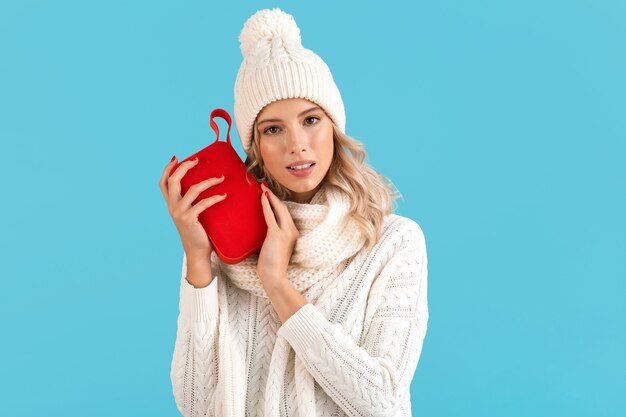 Elegante rubia sonriente hermosa joven sosteniendo altavoz inalámbrico escuchando música feliz vistiendo un suéter blanco y gorro de punto moda de estilo invernal posando aislado sobre fondo azul.