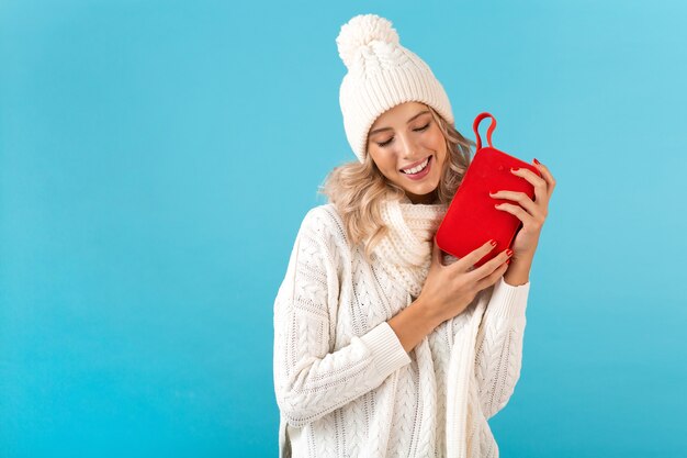 Elegante rubia sonriente hermosa joven sosteniendo altavoz inalámbrico escuchando música feliz vistiendo suéter blanco y sombrero de punto estilo de invierno posando de moda