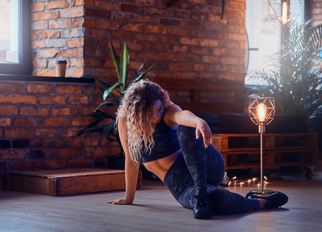 La elegante y sexy modelo de talla grande está haciendo yoga en un loft moderno. Lleva polainas deportivas, sujetador y zapatillas. La mujer tiene el pelo rubio y rizado. Pared de ladrillo y lámparas de fondo.