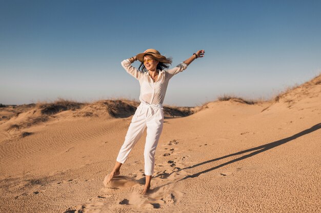 Elegante sonriente hermosa mujer feliz corriendo y saltando en la arena del desierto en traje blanco con sombrero de paja en la puesta de sol