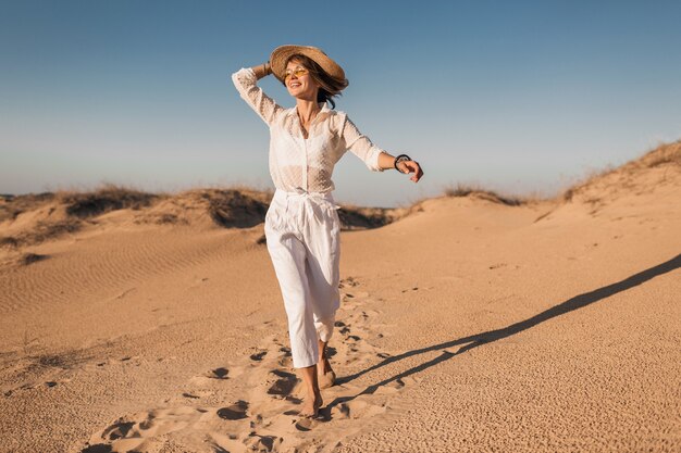 Elegante sonriente hermosa mujer feliz corriendo y saltando en la arena del desierto en traje blanco con sombrero de paja en la puesta de sol