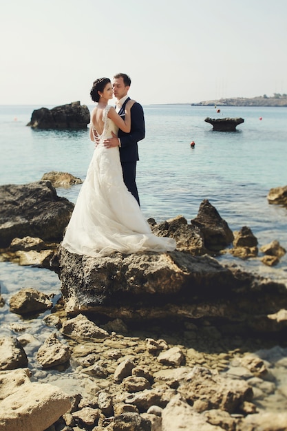 Elegante sonriente joven novia y el novio posando en las rocas en la playa