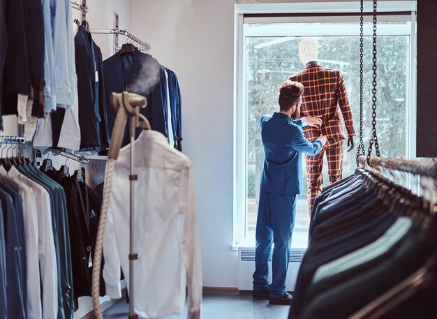 El elegante vendedor barbudo se preocupa por el traje en un maniquí en una tienda de ropa masculina.