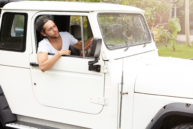 Elegante viajero caucásico con descanso durante el viaje de aventura de safari. Hombre joven con barba hipster en camiseta blanca sentado dentro de su automóvil SUV con tracción a las cuatro ruedas y mirando por la ventana abierta