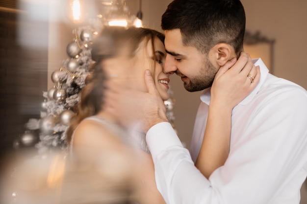 Foto gratuita elegantes hombres y mujeres vestidos con bata plateada se abrazan tiernamente de pie ante un árbol de navidad