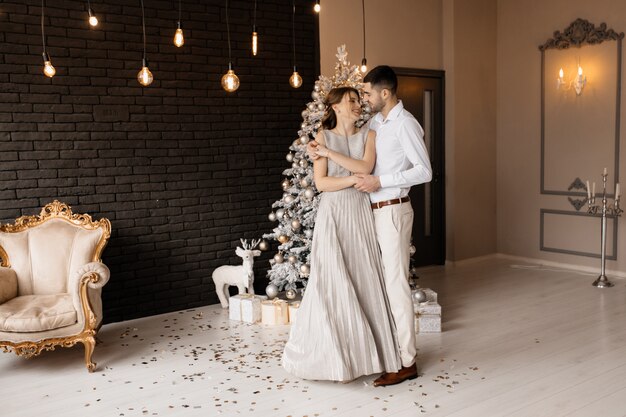 Elegantes hombres y mujeres vestidos con bata plateada se abrazan tiernamente de pie ante un árbol de Navidad