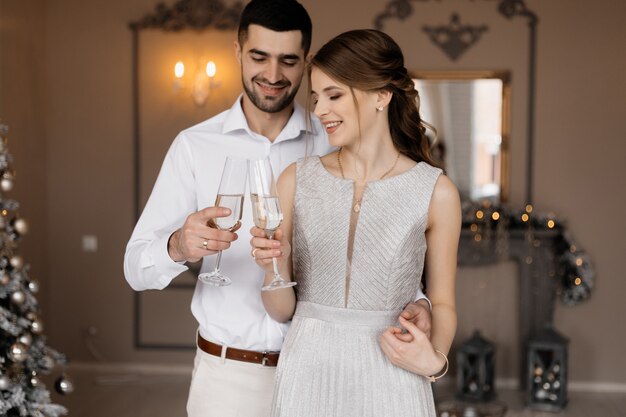 Elegantes hombres y mujeres vestidos con bata plateada se abrazan tiernamente de pie ante un árbol de Navidad