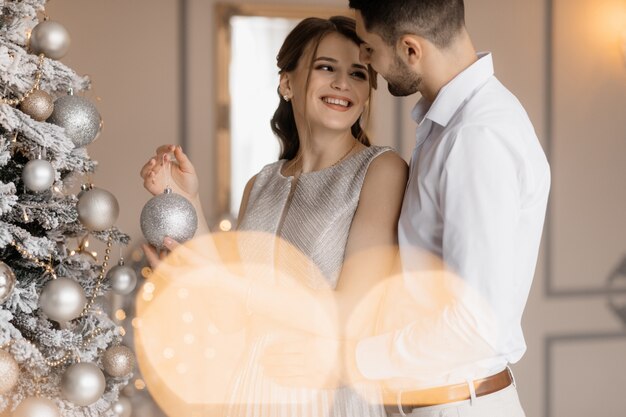 Elegantes hombres y mujeres vestidos con bata plateada se abrazan tiernamente de pie ante un árbol de Navidad