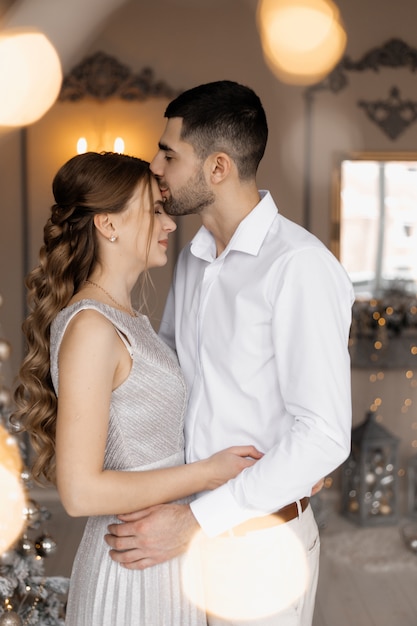 Elegantes hombres y mujeres vestidos con bata plateada se abrazan tiernamente de pie ante un árbol de Navidad