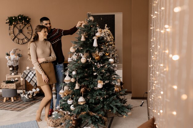 Elegantes hombres y mujeres vestidos con bata plateada se abrazan tiernamente de pie ante un árbol de Navidad