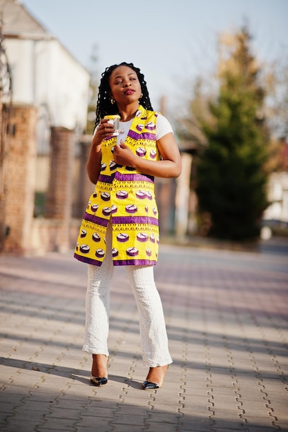 Elegantes mujeres afroamericanas con chaqueta amarilla posadas en la calle con bebida caliente en un vaso de papel desechable