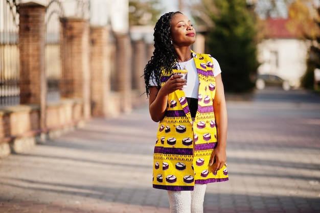 Elegantes mujeres afroamericanas con chaqueta amarilla posadas en la calle con bebida caliente en un vaso de papel desechable