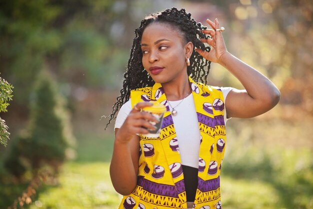 Elegantes mujeres afroamericanas con chaqueta amarilla posadas en la calle con bebida caliente en un vaso de papel desechable