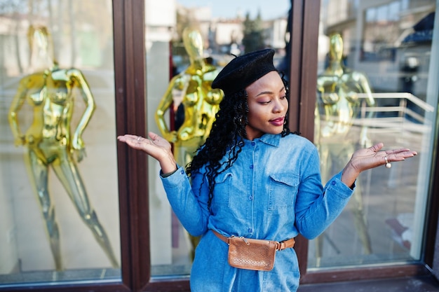 Elegantes mujeres afroamericanas de moda con jeans y boina negra contra un edificio moderno con tres maniquíes dorados