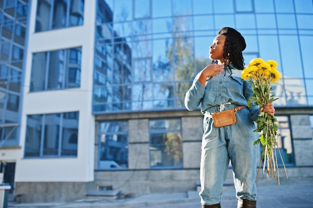 Elegantes mujeres afroamericanas de moda en jeans y boina negra con ramo de flores amarillas posadas al aire libre en un día soleado contra un edificio moderno azul