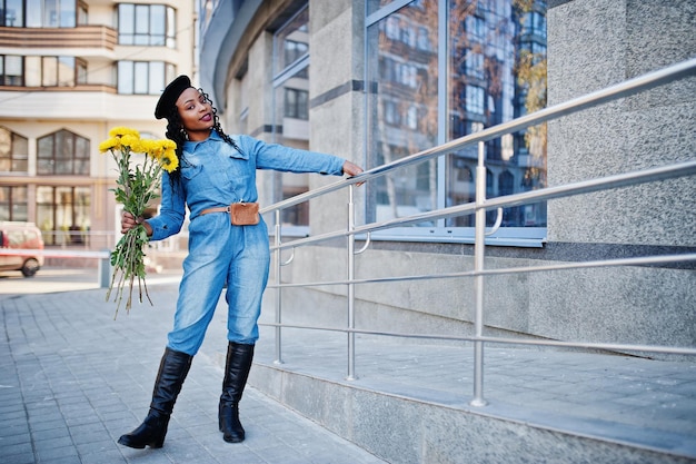 Elegantes mujeres afroamericanas de moda en jeans y boina negra con ramo de flores amarillas posadas al aire libre en un día soleado contra un edificio moderno azul