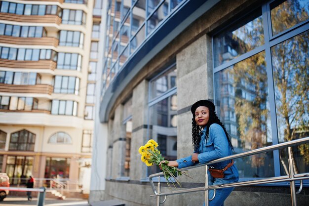 Elegantes mujeres afroamericanas de moda en jeans y boina negra con ramo de flores amarillas posadas al aire libre en un día soleado contra un edificio moderno azul