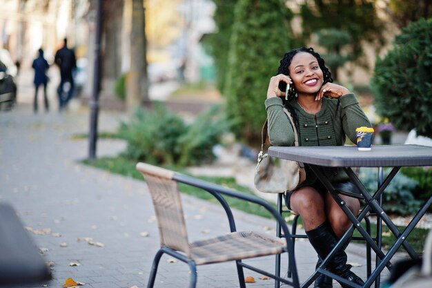 Elegantes mujeres afroamericanas de moda con suéter verde y falda negra posaron en un café al aire libre sentadas junto a la mesa con una taza de café y hablando por teléfono móvil