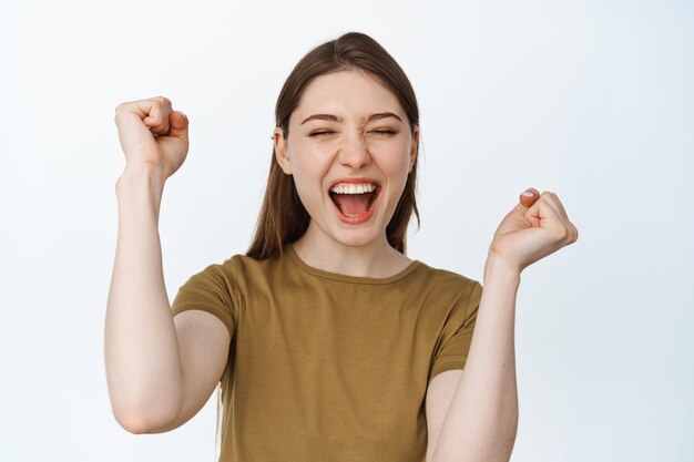 Emoción de ganar. Niña feliz animando y gritando de alegría, celebrando la victoria con las manos levantadas y la expresión de la cara complacida, triunfando en blanco.