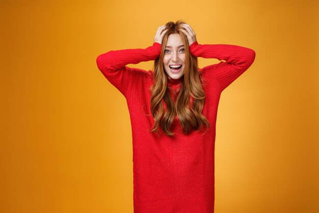 Emocionada y abrumada mujer encantadora pelirroja con vestido rojo de punto cálido gritando de emoción y felicidad tomados de la mano en el cabello sonriendo ampliamente impresionado con suerte inesperada sobre la pared naranja