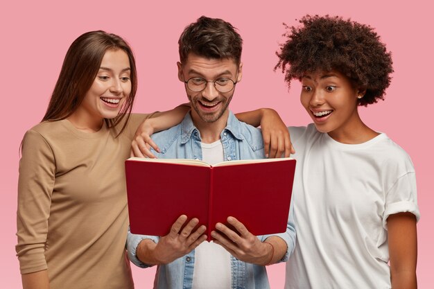 Emocionada alegre curiosa multiétnica dos mujeres jóvenes y chico guapo miran libros de texto, leen información, aislados sobre la pared rosa. Estudiantes interraciales felices aprenden algo antes del examen