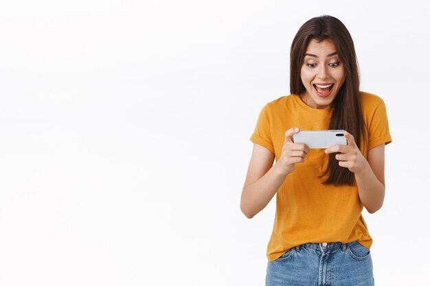 Emocionada, alegre y guapa mujer con camiseta amarilla jugando un nuevo e increíble juego móvil divertido, sosteniendo el teléfono inteligente horizontalmente, animando, sonriendo y mirando la pantalla del teléfono asombrada