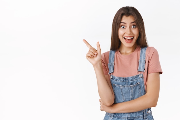 Emocionada alegre linda chica moderna en monos, camiseta apuntando hacia la esquina superior izquierda, sonriendo divertido discutir con la novia promoción increíble, sorteo, de pie fondo blanco emocionado