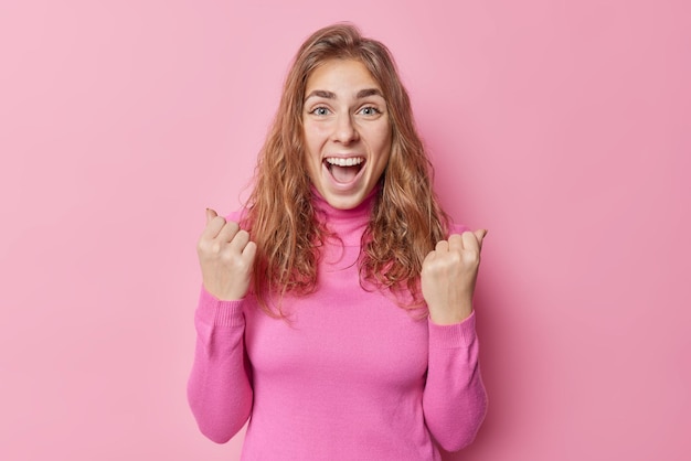 Emocionada y alegre mujer europea exclama que sí, aprieta los puños, triunfa sobre algo, celebra el éxito, usa un cuello de tortuga aislado sobre un fondo rosado, es solidario y feliz por algo