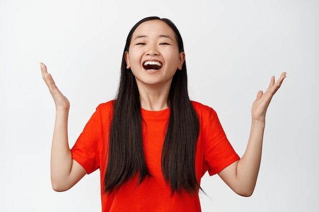 Emocionada y aliviada chica asiática extendió las manos de lado feliz riendo y sonriendo con pura alegría celebrando de pie sobre fondo blanco.