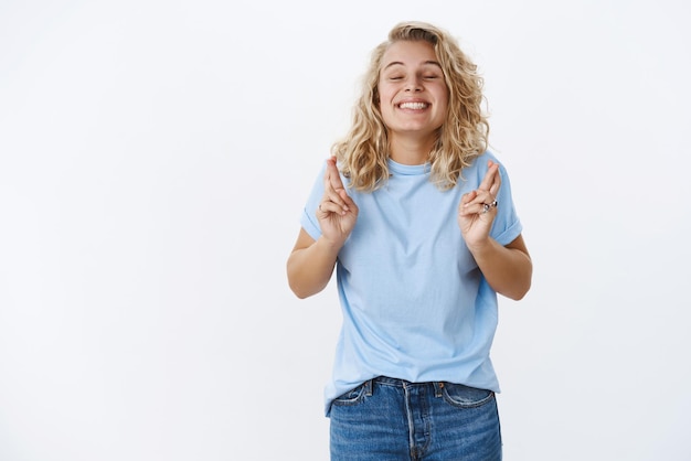 Emocionada anticipando a una linda chica rubia pidiendo un deseo con una dulce sonrisa optimista y los ojos cerrados cruzando los dedos para tener buena suerte mientras reza el sueño de esperanza hecho realidad siendo optimista sobre el fondo blanco