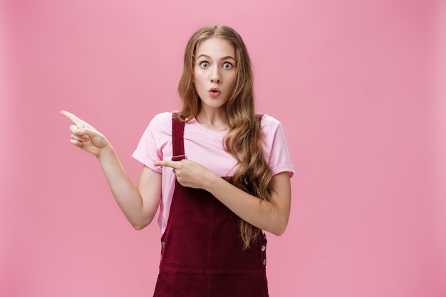 Emocionada, aturdida y sorprendida joven encantadora con maquillaje y peinado con curvas en un moderno peto de pana apuntando hacia la izquierda con los labios doblados por el interés y el asombro por ser curioso lo que sucedió.