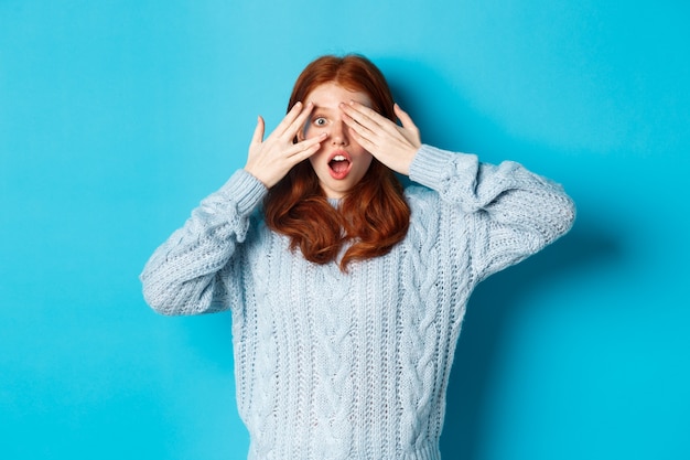 Emocionada chica adolescente pelirroja abre los ojos para ver la sorpresa navideña, recibe regalos, mira asombrada a la cámara, de pie sobre fondo azul.