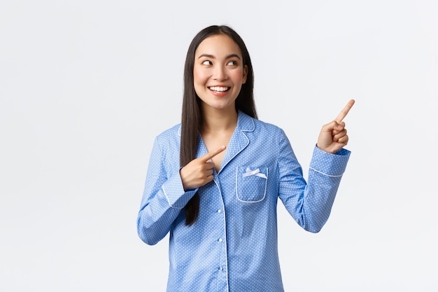 Emocionada chica bastante asiática en pijama azul apuntando con el dedo en la esquina superior derecha y mirando interesada. Mujer complacida en pijamas haciendo elección, mostrando publicidad genial, fondo blanco.