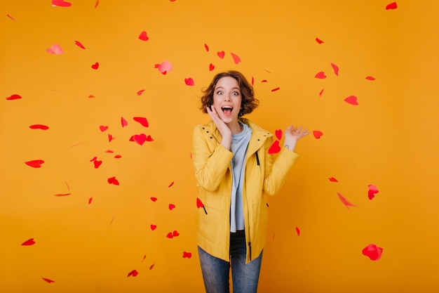 Emocionada chica blanca con cabello ondulado castaño claro posando en el día de San Valentín. Foto interior de mujer joven feliz de pie bajo corazones caídos y riendo.