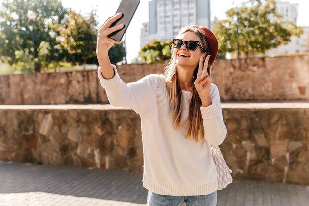 Emocionada chica francesa con selfie haciendo peinado largo. Foto al aire libre de magnífica dama riendo posando con smartphone.