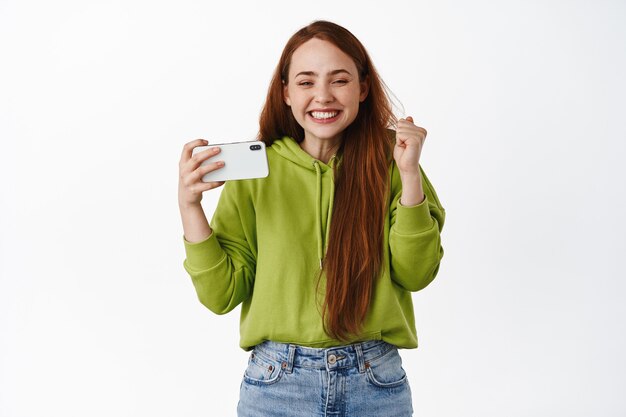 Emocionada chica de jengibre sosteniendo un teléfono inteligente horizontal y ganando, sonriendo complacida, triunfando de los logros en línea, de pie en blanco