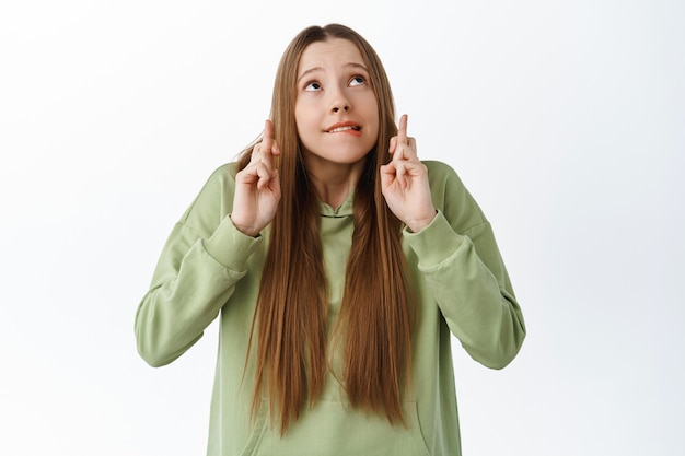 Emocionada chica linda mordiéndose el labio nerviosa, cruza los dedos y mira copyspace, esperando resultados con expresión preocupada, anticipando noticias, rezando, de pie sobre una pared blanca