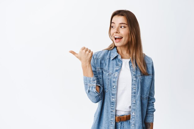 Emocionada chica milenaria gire la cabeza y señale con el pulgar hacia la izquierda, revisando una buena oferta promocional, demostrando publicidad en el espacio de la copia, blanco