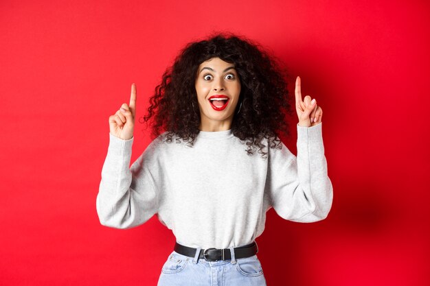 Emocionada chica moderna mostrando promo apuntando con el dedo hacia arriba y sonriendo sorprendido contando grandes noticias de pie ...