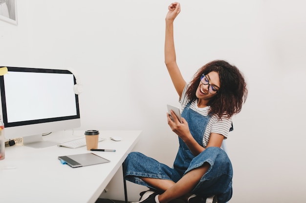 Emocionada chica negra sentada con las piernas cruzadas y riendo feliz, leyendo mensajes telefónicos