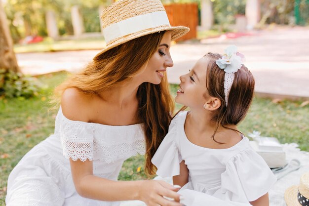 Emocionada chica de pelo castaño con cinta mirando a madre sonriente con maquillaje elegante. Retrato al aire libre de mujer alegre con sombrero y vestido blanco, pasar tiempo libre con su hija en el parque durante el fin de semana.