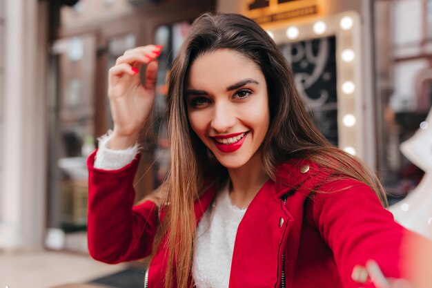 Emocionada chica de pelo castaño con maquillaje elegante haciendo selfie en espacio urbano borroso