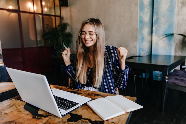 Emocionada chica de pelo largo que se divierte durante el trabajo con la computadora. Foto interior de freelance femenino sonriente usando laptop en acogedor café.