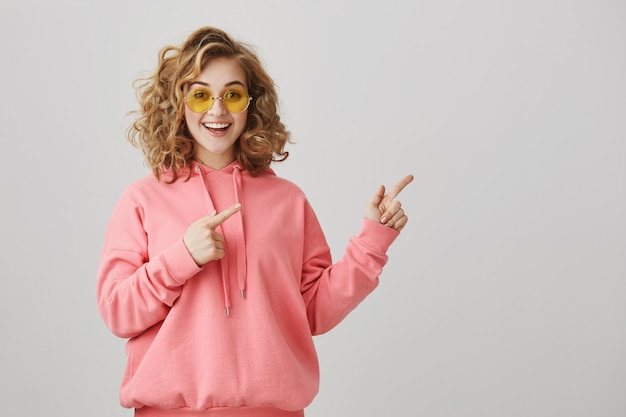 Emocionada chica de pelo rizado con estilo en gafas de sol apuntando a la derecha, mostrando el camino