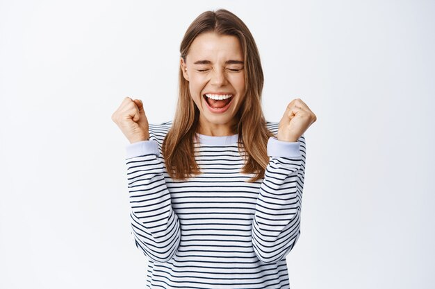 Emocionada chica rubia gritando de alegría, saltando de la felicidad y celebrando la victoria, sonriendo asombrada, triunfando del éxito, pared blanca