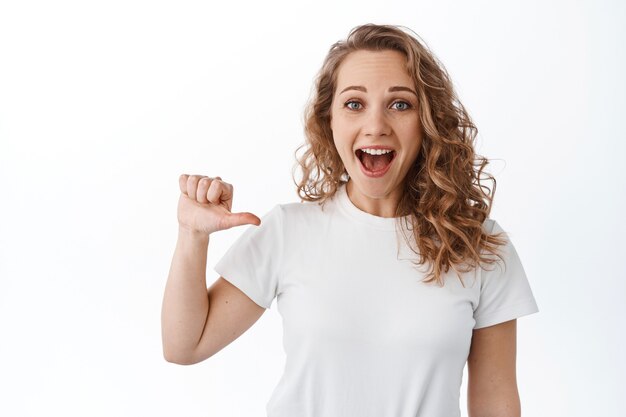 Emocionada chica rubia jadeando asombrada, señalando a sí misma con una sonrisa alegre y optimista, mostrándola, autopromocionando, de pie sobre una pared blanca