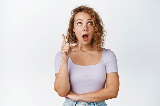 Emocionada chica rubia con la mandíbula caída, apuntando y mirando hacia arriba con rostro fascinado, revisando la oferta promocional, el anuncio, de pie en blanco