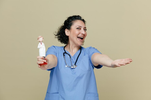 Emocionada doctora de mediana edad con uniforme y estetoscopio alrededor del cuello mirando a la cámara estirando la mano y la figura del doctor hacia la cámara aislada en el fondo verde oliva